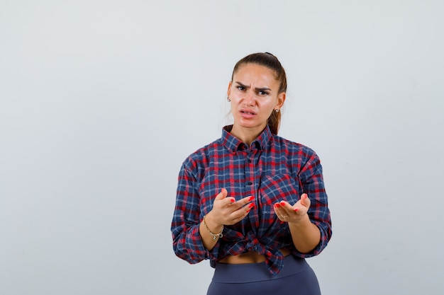 Giovane donna che mostra gesto impotente in camicia a scacchi, pantaloni e sembra esitante. vista frontale.