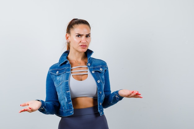 Young female showing helpless gesture in crop top, jacket, pants and looking serious. front view.