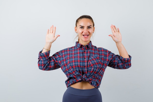 Young female showing palms in surrender gesture in checkered shirt, pants and looking happy , front view.