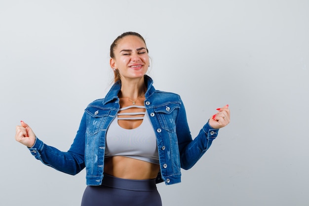 Young female showing winner gesture in crop top, jacket, pants and looking blissful. front view.