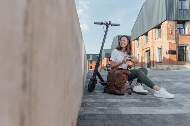 Young female sitting on urban stairs and browsing smart watch on electric scooter