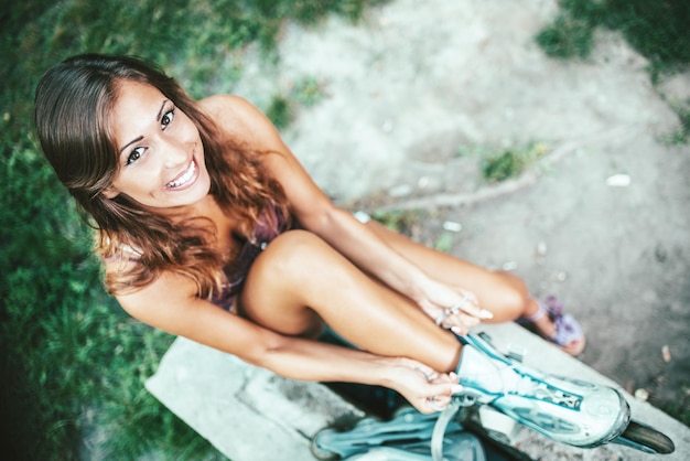 Young female skater is sitting in the park and ties her rollers preparing for roller skating in the city park.