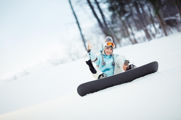 Young female snowboarder sitting on slope and showing thumb up