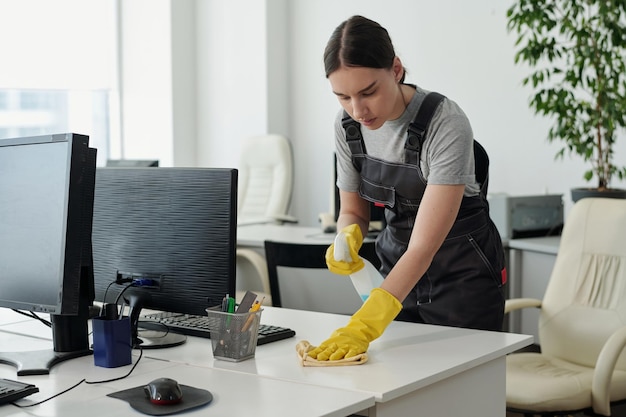 young female staff of cleaning service company wiping desk in openspace office