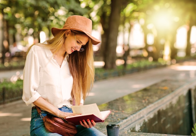 Молодая студентка, одетая в одежду повседневного стиля, читая книгу, сидя в городе на открытом воздухе.