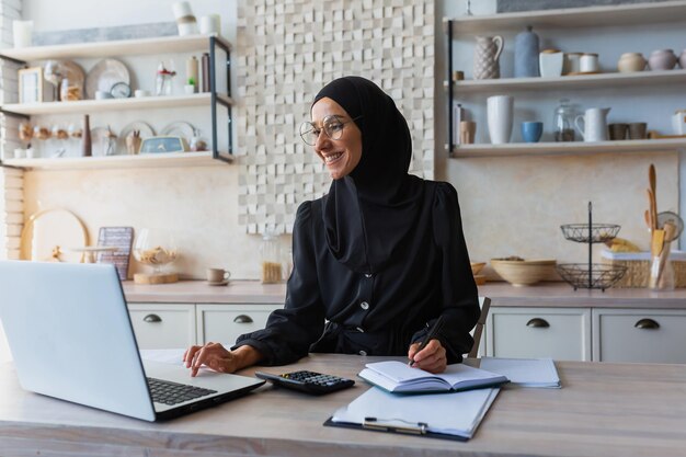 A young female student of oriental origin in a hijab sits at home in the kitchen with a laptop and