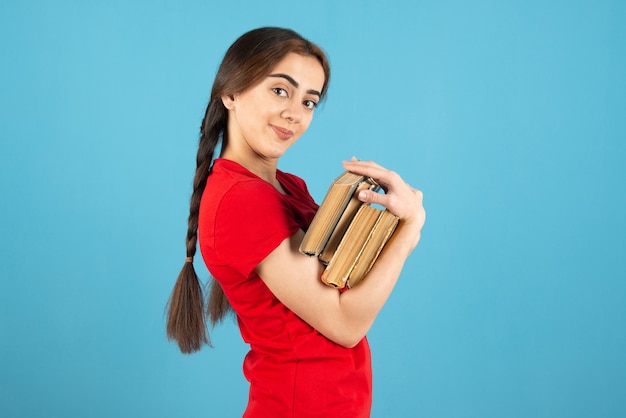 Young female student in red t-shirt with books standing on blue wall.