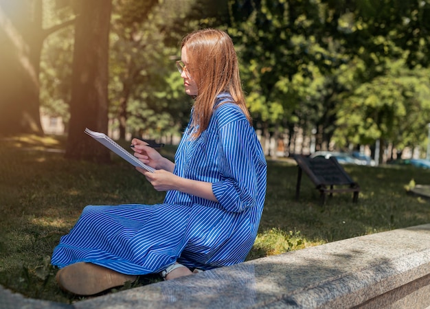 Giovane studentessa seduta sull'erba con blocco note o libro accademico e imparare a leggere e scrivere ...