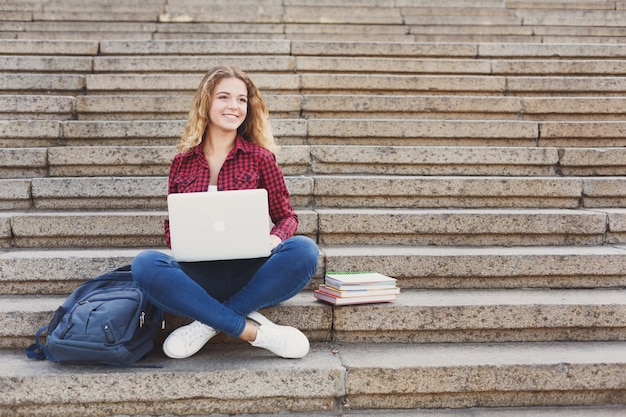 Молодая студентка сидит на лестнице на улице и работает с ноутбуком, готовится к экзаменам или отдыхает в университетском городке. Технологии, образование и концепция удаленной работы, пространство для копирования