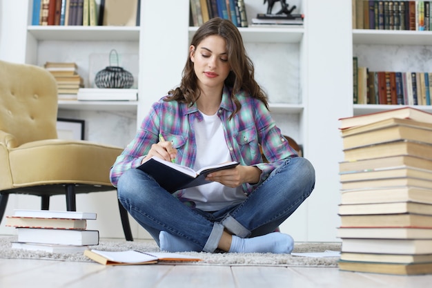 Young female student studying at home, writing article for publication, sitting on floor against cozy domestic interior, surrounded with pile of books.