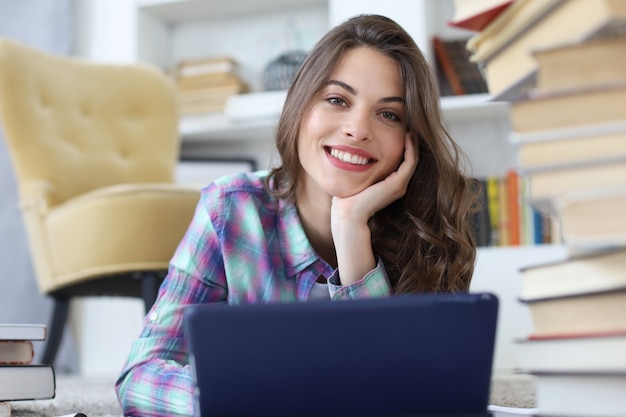 Young female student studying at home, writing article for publication, sitting on floor using tablet against cozy domestic interior, surrounded with pile of books.