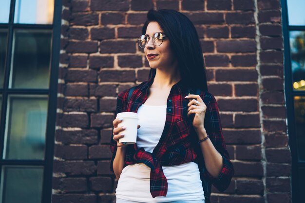 Young female student in stylish spectacles enjoying recreation outdoors with delicious coffee