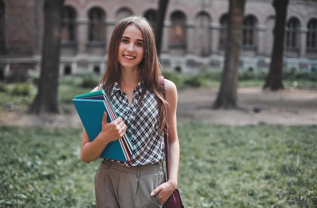 Young female student with multi-colored notebooks near the campus and smiling