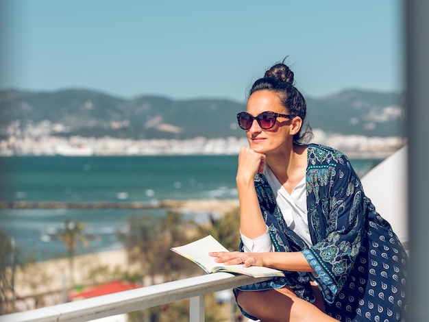 Молодая женщина в летней одежде и солнцезащитных очках сидит на балконе с книгой, наслаждаясь видом на море в солнечный день