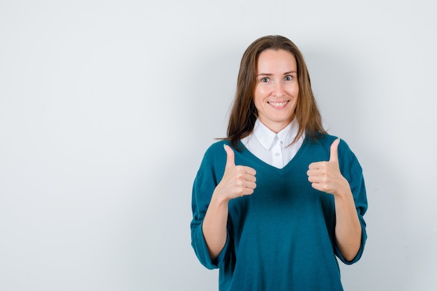 Young female in sweater over shirt showing double thumbs up and looking cheerful , front view.