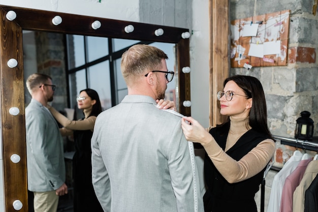 ワークショップやファッションスタジオで鏡のそばに立っている間、若い男性のジャケットの対策を取っている若い女性の仕立て屋