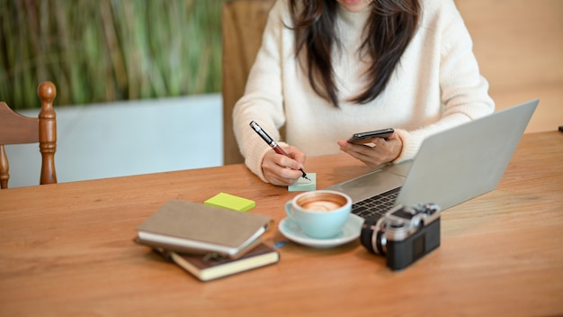 Young female taking notes on a sticky colourful notepaper while looking on smartphone sits at caf