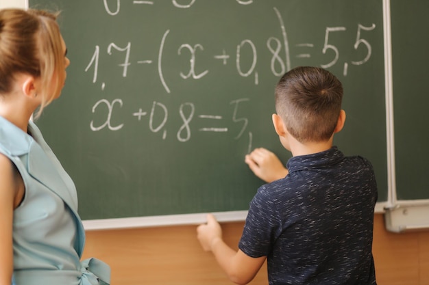Young female teacher help schoolboy boy stand by the blackboard