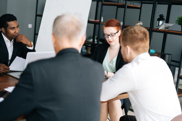 Young female team leader in glasses holding corporate meeting with multi-ethnic colleagues in boardroom sitting at desk. Serious businesswoman discussing project with business partners in office.