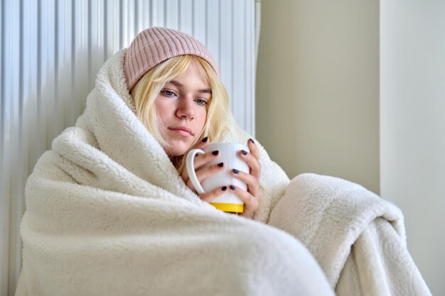Young female teenager warming herself with blanket heating radiator hot tea