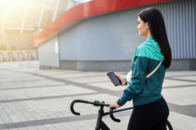 Young female touching the handlebars of a bicycle and using phone