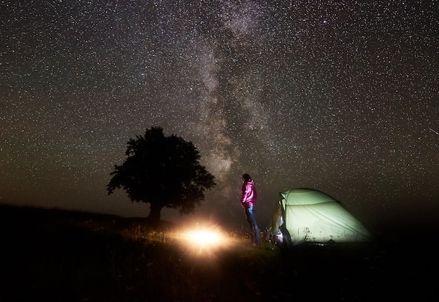 Foto giovane turista femminile che riposa vicino alla tenda illuminata, accampandosi in montagna di notte sotto il cielo stellato