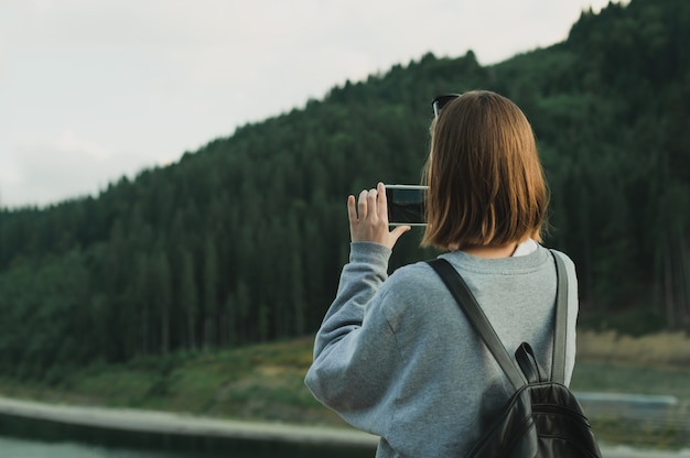 Young female traveler in grey sweatshirt taking photo on smartphone of mountain forest landscape