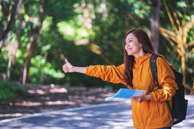 산길에서 엄지손가락으로 차를 멈추고 히치하이킹하는 젊은 여성 여행자