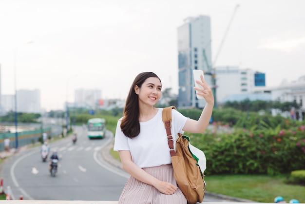 街の中心部の橋に座って携帯電話で自分撮り写真を作る若い女性旅行者