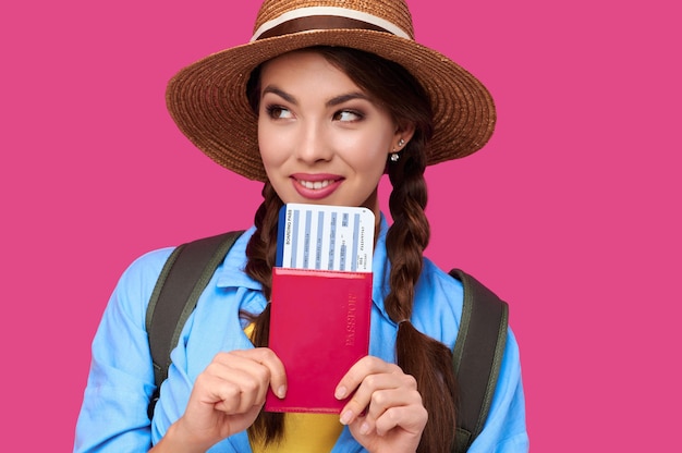 Young female traveler wearing casual clothes and straw hat holding passport with ticket on pink isolate background. Tourism concept. Studio shot