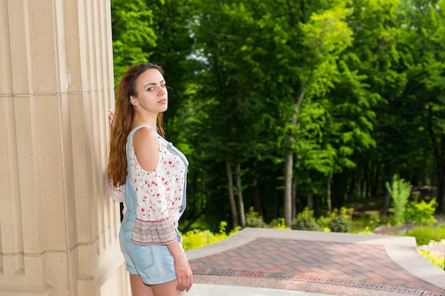 Young female turned around to look into the camera standing near stone wall outside facing trees in park or yard