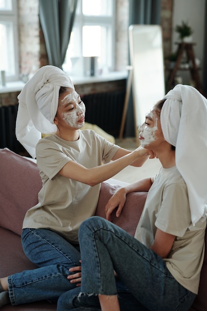 Young female twins with facial clay mask sitting on couch and relaxing after bath while one of them helping her sister put towel on head