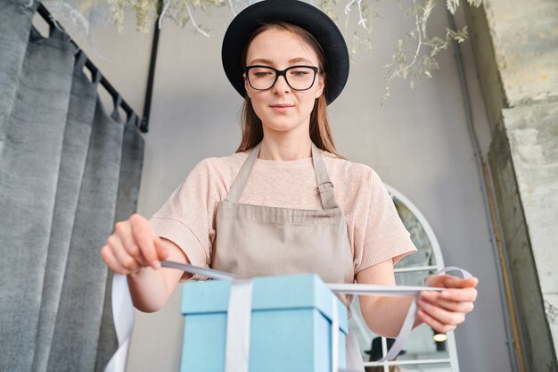 Young female tying up silk ribbon on top of blue giftbox while working in studio or shop