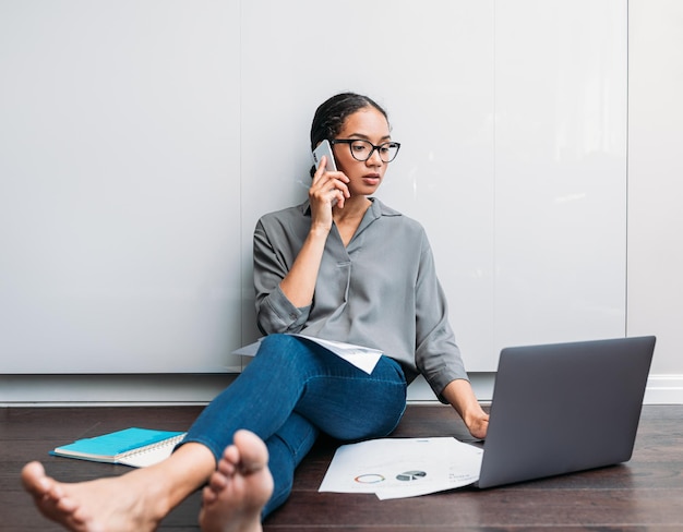 Young female using a laptop and talking on a cell phone while sitting on the floor at home