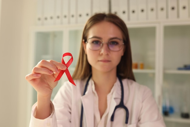 Young female virologist in glasses holds red ribbon as symbol of AIDS disease in hospital