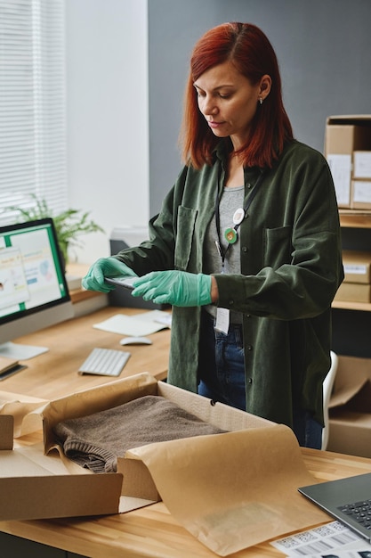 Young female volunteer with smartphone taking photo of folded sweater in box