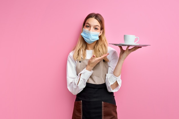 Young female waiter offering cup of coffee isolated on pink wall