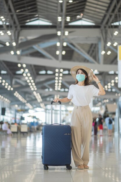 마스크를 쓴 젊은 여성 공항 보호용 가방을 들고 걸어다니는 코로나바이러스 질병 감염 모자를 쓴 아시아 여성 여행자 백신 부스터 용량 개념 후 여행할 시간