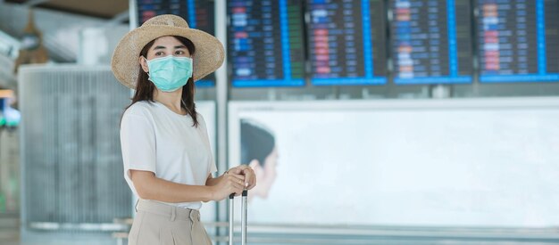 空港保護で荷物を持って歩いているフェイスマスクを身に着けている若い女性コロナウイルス病感染症帽子をかぶったアジアの女性旅行者旅行する時間テストアンドゴーとワクチンブースター用量の概念