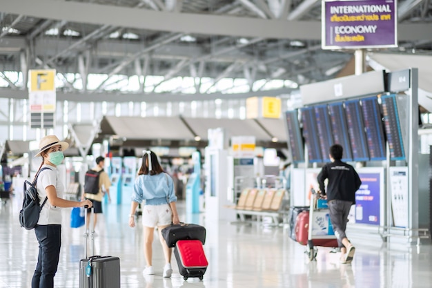 공항 터미널, 보호 코로나 바이러스 질병 (Covid-19) 감염, 아시아 여성 여행자 모자와 함께 걷는 짐과 얼굴 마스크를 착용하는 젊은 여성. 새로운 일반 및 여행 거품 개념