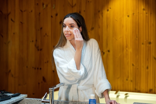 白いバスローブを着た若い女性が、バスルームでの毎日の美容処置中にコットンパッドと化粧品で顔の皮膚を拭きます