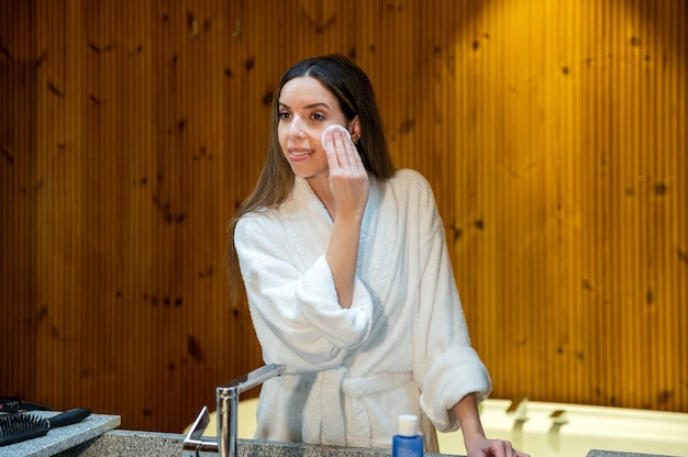 Giovane donna in accappatoio bianco che pulisce la pelle del viso con un batuffolo di cotone e un prodotto cosmetico durante la procedura di bellezza quotidiana in bagno