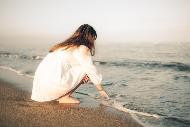 Young female in a white dress touching the water on a seashore