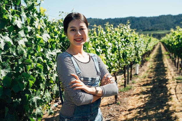 Young female winemaker in vineyard with arms crossed face camera smile confidently. asian lady standing in her working place in grape winery. beautiful farmer in overalls in the farm in spring.