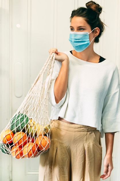 A young female with a face mask and a reusable mesh shopping bag full of fruits and vegetables