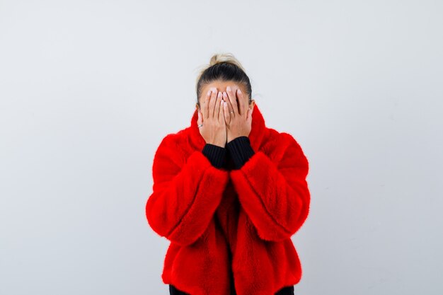 Young female with hands on face in sweater, red fur coat and looking scared , front view.