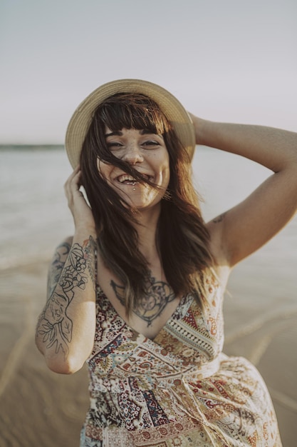 Young female with tattoos wearing a dress and straw hat on blurred ocean surface