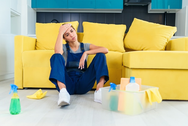 Young female worker of the cleaning service sitting near the couch and feeling tired after washing floor on cuisine.