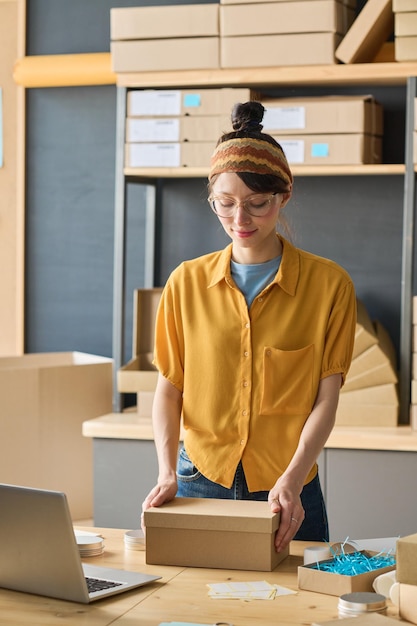 Young female worker packing goods in cadboard boxes at table in warehouse before delivery