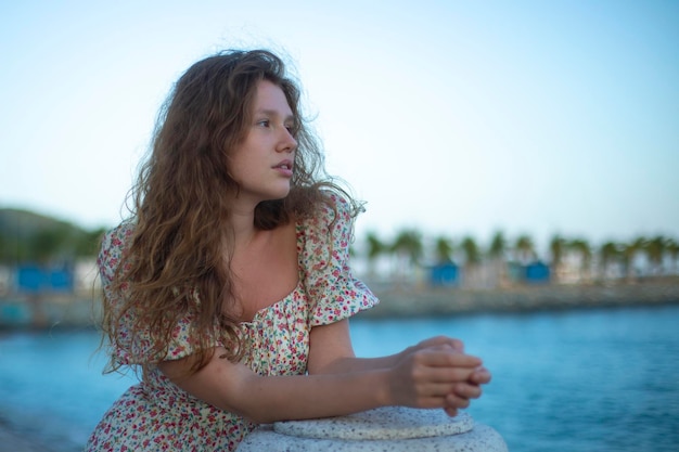 Young feminine girl in a dress stands on promenade by sea and enjoys a warm summer day